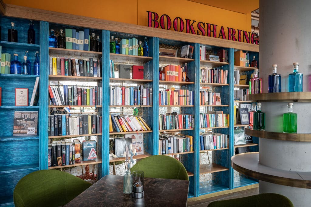 Regal mit einer großen Auswahl an Büchern im Booksharing-Bereich des Brucks Café und Restaurant im Brucklyn Quartier, mit blau gestrichenem Holz und dekorativen Flaschen
